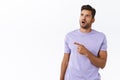 Astonished and speechless surprised handsome hipster guy in purple t-shirt, drop jaw and gasping amazed, pointing