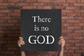 Atheism. Man holding sign with text There Is No God near brick wall, closeup