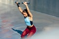 An athelte young woman doing exercises with dumbells outdoors on a sunny day at grey background. Fit female squats with dumbbells