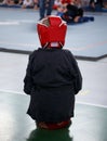 A young athlete boy in boxing gloves and a protective headgear looks at the tatami with the fighters.