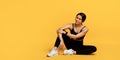 Athletic african american lady resting after workout, sitting on floor at yellow background and looking at empty space