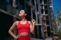 Athletic cheerful young woman in red sports top and leggins touching her ponytail and enjoying sunlight. Blurred city