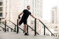 Athletic man stretching legs before workout outdoors. Runner in black sportswear excercising at morning holding handrail Royalty Free Stock Photo