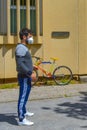 A young guy with white face mask for covid protection stands in front of building, colorful bicycle in background. Sportswear guy