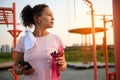 Athletic woman with smartphone and bottle of water resting after physical exercises on a sports ground with exercise equipment and
