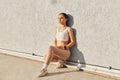 Athletic young woman sitting and leaning against gray wall looking away with calm and serious facial expression, wearing stylish Royalty Free Stock Photo