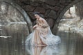 Atmospheric outdoor portrait of sensual young woman wearing elegant dress sitting under the bridge of river.