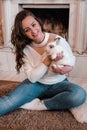 Atractive young woman sitting next to the fireplace and holding rabbit. Pets and people. Home sweet home, Hygge. Animals