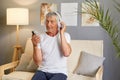 Attentive older gentleman wearing white T-shirt sitting on cough holding his mobile phone and headphones listening to music or Royalty Free Stock Photo