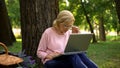 Attentive senior lady sitting in park with laptop and working online, freelance