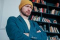 Attractive adult bearded angry man hipster in yellow hat looks at camera and furrowed his brows on books wall background