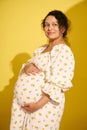 Attractive African American gravid woman, gently holding her belly, smiling looking aside, isolated yellow background Royalty Free Stock Photo