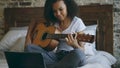 Attractive african american teenager girl concentraing learning to play guitar using laptop computer sitting on bed at Royalty Free Stock Photo