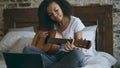 Attractive african american teenager girl concentraing learning to play guitar using laptop computer sitting on bed at Royalty Free Stock Photo