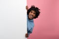 Attractive African American teenager hiding behind blank poster with copy space on pink studio background, mockup