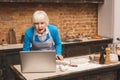 Attractive amazed shocked senior aged woman is cooking on kitchen. Grandmother making tasty baking. Using laptop