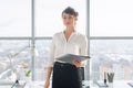 Attractive ambitious businesswoman standing in modern office, holding paper folder, looking at camera, smiling. Royalty Free Stock Photo