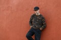 Attractive American young man in a camouflage shirt in jeans in a fashionable black cap posing standing near a vintage building Royalty Free Stock Photo