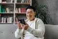 Attractive asian man in casual wear sitting on comfy couch with modern smartphone in hands. Royalty Free Stock Photo