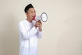 attractive Asian muslim man in white shirt with skullcap speaking louder using megaphone, promoting product.