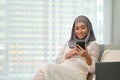 Attractive Asian Muslim pregnant woman resting, using her mobile phone on the sofa Royalty Free Stock Photo