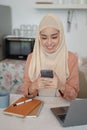 An attractive Asian Muslim woman wearing a hijab is using her smartphone in the kitchen Royalty Free Stock Photo