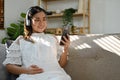 Attractive Asian pregnant woman wearing headphones, listening to music while sitting on sofa Royalty Free Stock Photo