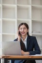 Attractive Asian woman using laptop in online meeting. Businesswoman using mobile phone talking with colleague sitting Royalty Free Stock Photo