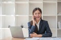 Attractive Asian woman using laptop in online meeting. Businesswoman using mobile phone talking with colleague sitting Royalty Free Stock Photo