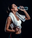 Attractive athletic young woman with perfect body drinking water from a bottle with towel around her neck against black background Royalty Free Stock Photo