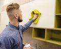 Attractive bearded man cleaning the handleless cabinet doors with a yellow rag in a modern kitchen