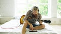 Attractive bearded man sitting on bed learning to play guitar using tablet computer in modern bedroom at home Royalty Free Stock Photo