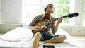 Attractive bearded man sitting on bed learning to play guitar using tablet computer in modern bedroom at home Royalty Free Stock Photo