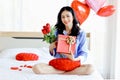 Attractive beautiful Asian woman holding red rose bouquet flower and gift box while sitting on white bed in bedroom, smiling girl Royalty Free Stock Photo