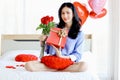 Attractive beautiful Asian woman holding red rose bouquet flower and gift box while sitting on white bed in bedroom, smiling girl Royalty Free Stock Photo