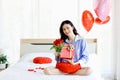 Attractive beautiful Asian woman holding red rose bouquet flower and gift box while sitting on white bed in bedroom, smiling girl Royalty Free Stock Photo