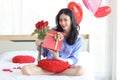 Attractive beautiful Asian woman holding red rose bouquet flower and gift box while sitting on white bed in bedroom, smiling girl Royalty Free Stock Photo