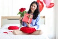 Attractive beautiful Asian woman holding red rose bouquet flower and gift box while sitting on white bed in bedroom, smiling girl Royalty Free Stock Photo