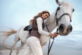 Attractive and beautiful young woman wearing stylish jockey outfit is holding the reins and posing with the white horse