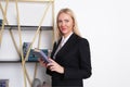 Attractive blonde business woman in business suit stands at bookshelf and chooses a book to read