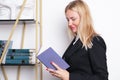 Attractive blonde business woman in a business suit stands at the bookshelf and reads a book