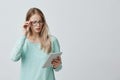 Attractive blonde female manager in stylsh eyewear works, look nervously at screen of tablet finds out unpleasant news Royalty Free Stock Photo