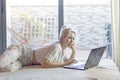 Beautiful young woman relaxing on her bed in a large casual shirt working with her laptop