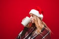 Attractive blonde girl in Christmas hat sings with closed eyes hot drink from big cup on red background, wears warm plaid. Winter