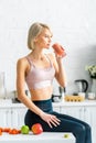 Blonde girl in sportswear drinking tasty smoothie while sitting in kitchen near apples