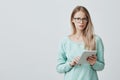 Attractive blonde office worker in stylsh eyewear works, look at camera with appeal, holds tablet in hands, surfs
