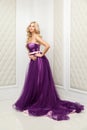 Attractive blonde woman in fashionable purple classic evening dress with makeup and long wavy hairstyle standing and posing near Royalty Free Stock Photo