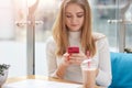 Attractive blonde woman in white shirt sitting at cafe on blue sofa, holding mobile phone, chatting with her friends using wi fi, Royalty Free Stock Photo