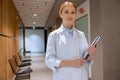 Attractive blonde young female doctor standing in hospital corridor and holding clipboard. Royalty Free Stock Photo