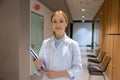 Attractive blonde young female doctor standing in hospital corridor and holding clipboard. Royalty Free Stock Photo